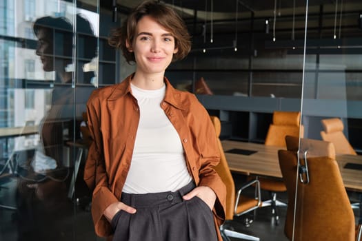 Portrait of young, self-assured woman standing in office, manager in her workplace, holding hands in pockets of trousers.