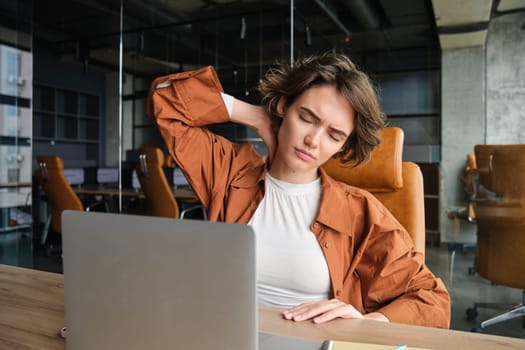 Portrait of woman touching her neck, massaging her tensed muscles, feels pain after sitting and working all day with laptop, works in office.