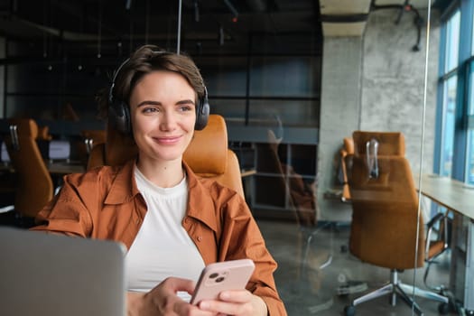 Close up portrait of beautiful smiling woman working in office, wearing headphones, sitting in front of laptop, holding smartphone.