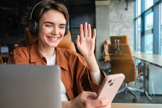 Portrait of working office woman sitting with laptop, joins video chat, records video for company social media in wireless headphones, waves hand at smartphone app.