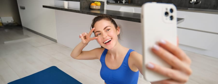 Close up portrait of beautiful brunette sportswoman, taking selfie on smartphone, posing on fitness mat with mobile phone, records her fitness exercises, workout training.