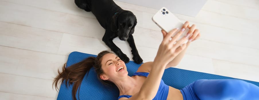 Portrait of happy young fitness woman, lying on mat and taking selfie with a dog in living room, workout with her pet.