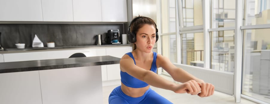 Image of young woman doing aerobics workout form home, listening music in wireless headphones, making squats exercises, fitness in living room.