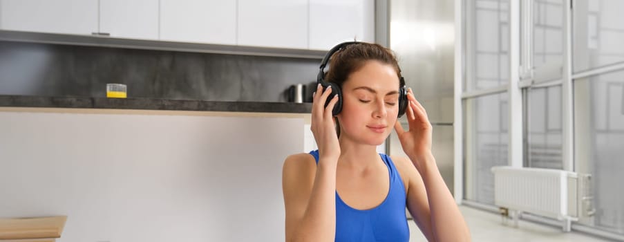Young woman puts on wireless headphones to listen music during workout at home, wears sportsbra, smiles with pleased face.
