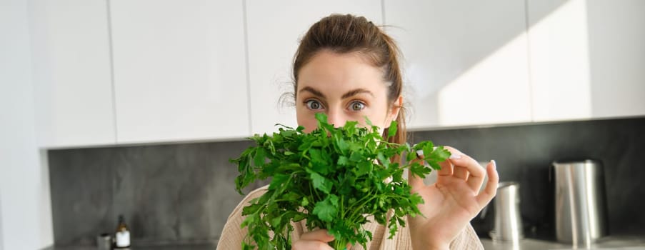 Portrait of beautiful woman cooking, holding fresh parsley, adding herbs while making salad, healthy meal in the kitchen.