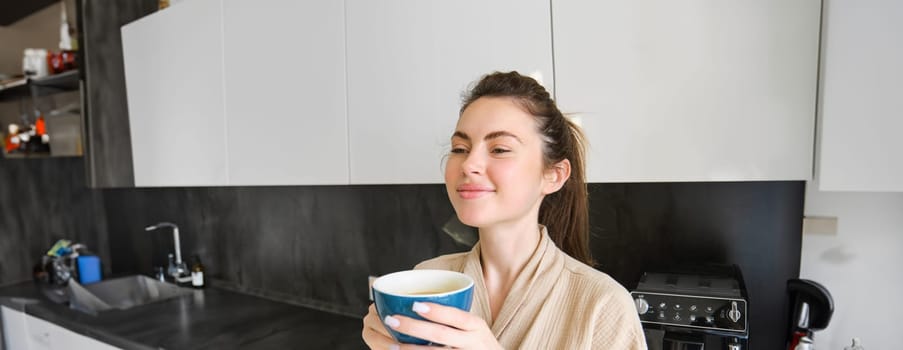 Portrait of smiling woman drinks coffee, stands in the kitchen and enjoys delicious cup of cappuccino in the morning, looks happy at camera.