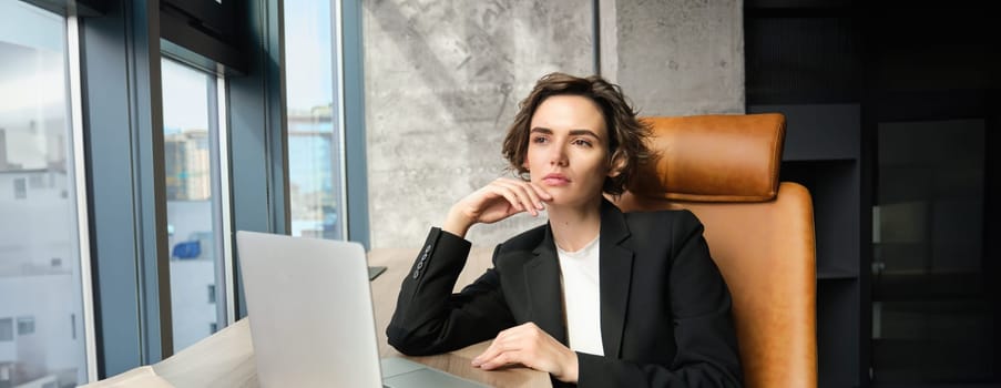 Portrait of confident businesswoman in office, sitting with laptop, looking serious outside window, thinking, solving issue at work.