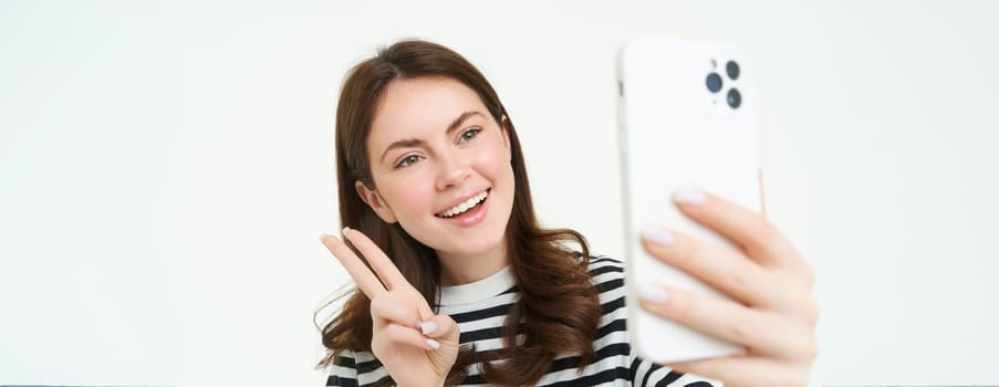 Portrait of young woman taking selfies on smartphone, posing for photo,. using mobile phone app for taking funny and cute pictures, isolated on white background.