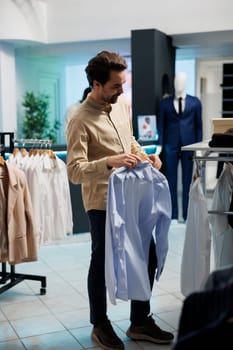 Shopper standing near clothes racks in fashionable boutique and holding shirt on hanger. Young man buyer choosing trendy formal wear outfit while shopping in mall showroom