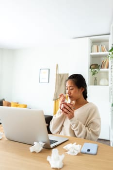 Young Chinese woman feeling sick working from home office. Asian female blowing nose with tissue holding cup of healing herbal tea. Vertical image. Wellness concept.