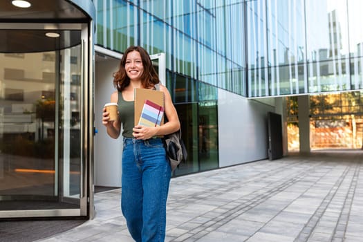 Young redhead happy female university student with backpack walking to class in college campus holding cup of coffee and notebooks. Copy space. Higher education concept.