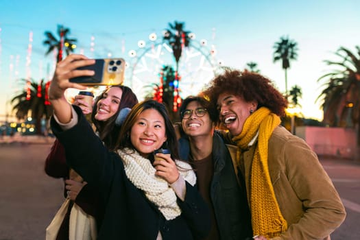 Happy multiracial group of smiling and cheerful college friends taking selfie together in the city using mobile phone. Friends having fun in Christmas winter market outdoors. Technology and lifestyle concept.
