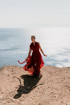 Side view a Young beautiful sensual woman in a red long dress posing on a rock high above the sea during sunrise. Girl on the nature on blue sky background. Fashion photo.