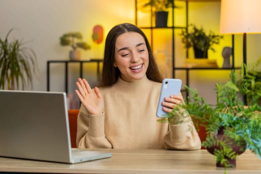 Caucasian young woman taking selfie on smartphone waving hello communicating video call online with subscribers, recording stories vlog for social media. Female girl sitting at home office at table