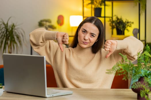 Dislike. Upset unhappy Caucasian woman showing thumbs down hands sign gesture expressing discontent disapproval dissatisfied bad work at home office indoors. Displeased young girl in room at table