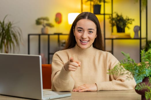 Hey you. Happy satisfied Caucasian young woman smiling excitedly and pointing to camera choosing lucky winner indicating to awesome you inviting approve. Female girl sitting at home office at table