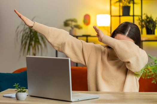 Trendy cheerful positive Caucasian young woman having fun dancing, dabbing raising hands, making dub winner dance gesture celebrating victory triumph. Happy female girl sitting at home office at table