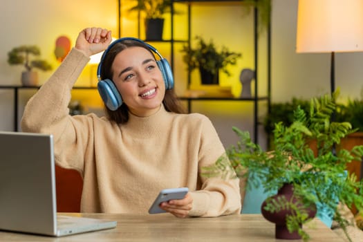 Happy relaxed overjoyed Caucasian young woman in wireless blue headphones sits at home table choosing listening favorite disco music in smartphone dancing entertaining. People weekend activities