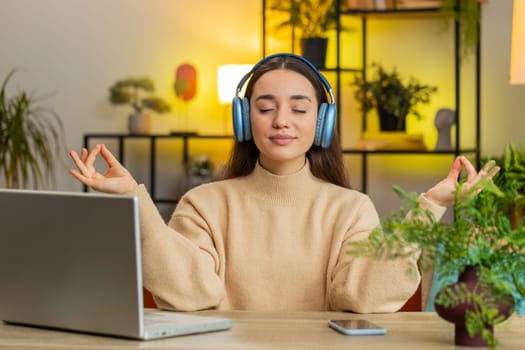 Keep calm down, relax. Pretty young woman listening music breathes deeply, eyes closed meditating with concentrated thoughts, peaceful mind. Young Caucasian girl sits at home table in room apartment