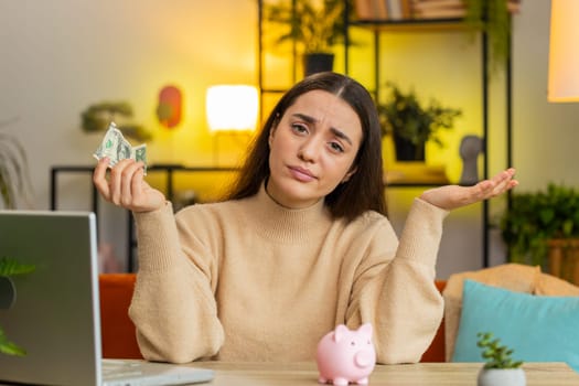 Poor Caucasian young woman insufficient amount of money, holding piggybank and one dollar banknote. Financial crisis. Bankruptcy. Poverty and destitution. Female girl sitting at home room at table