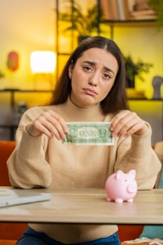 Poor young woman insufficient amount of money, holding piggybank and one dollar banknote. Financial crisis. Bankruptcy. Poverty and destitution. Female girl sitting at home office at table. Vertical