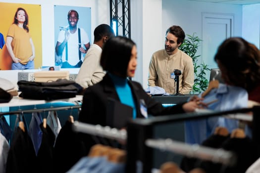 Clothing store cashier at clothing store engaging in conversation with customer at counter. Buyer waiting in line while man employee scanning apparel and talking with client