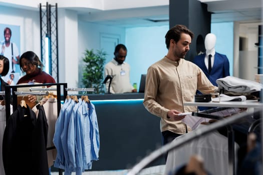 Caucasian man browsing through rack of clothing items at store, searching for casual shirt. Fashion boutique customer selecting size while exploring outfit in shopping mall showroom