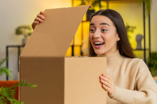 Happy Caucasian young woman unpacking delivery parcel sitting at home office room. Smiling satisfied girl shopper online shop customer opening cardboard box receiving purchase gift by postal shipping