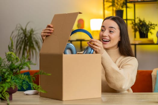 Happy Caucasian young woman unpacking delivery parcel sitting at home. Smiling satisfied girl shopper, online shop customer opening cardboard box receiving purchase gift headphones by postal shipping
