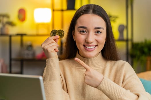Happy Caucasian woman freelancer successful developer programmer holding two gold coins. Girl stock trader earning bitcoins after online monitoring trading operations sitting at home office at table