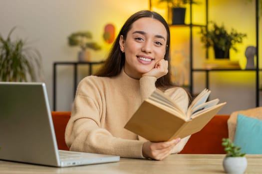 Caucasian smiling woman reading interesting book, turning pages smiling enjoying literature, taking a rest sits at table desk. Portrait of peaceful cheerful girl relaxing at home apartment living room
