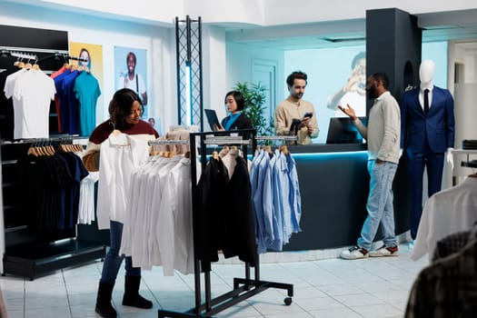 African american woman browsing rack with white shirts and selecting size in clothing store. Customer holding formal wear apparel and examining fit while shopping in fashion boutique