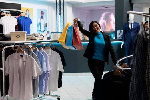 Smiling young asian woman holding paper bags while shopping in clothing store and looking at camera. Fashion boutique excited customer carrying packages and enjoying purchase