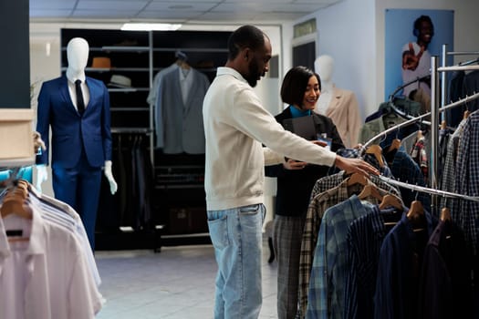 Boutique assistant helping african american man with clothing choice while browsing apparel rack. Customer checking casual plaid shirts on hangers and talking with store employee