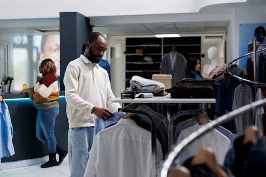 Young african american man shopping for casual apparel and browsing rack with shirts in menswear boutique. Clothing store customer choosing outfit and checking garment style