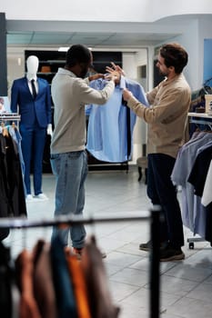 African american man selecting formal outfit and asking clothing store assistant for advice. Shopping mall boutique employee and customer discussing shirt and tie style match