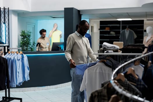 Clothing store client looking at casual shirt, examining style and fabric quality before buying. African american man browsing through apparel rack while shopping in fashion boutique