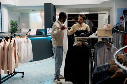 Clothing store customer holding shirt to body and asking fashion consultant for advice. African american man trying on formal apparel while shopping and getting help from boutique worker