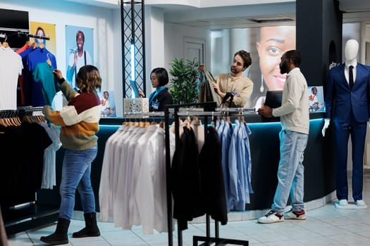 Cashier and customer interacting at clothing store counter desk. African american man buying casual trendy apparel and standing at cash register in shopping mall boutique