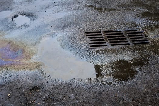 Oil or gasoline stains caught in a puddle against the background of an old asphalt surface with a grate flowing into a storm drain. Environmental problems of water pollution.