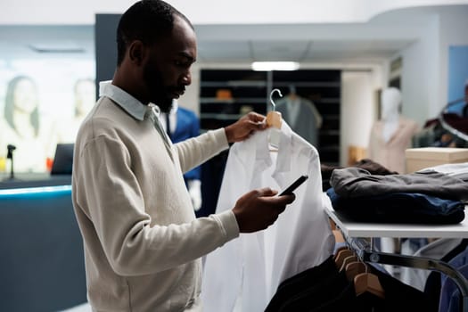 Clothing store client selecting formal outfit from hanging rack and checking price on mobile website. African american man holding shirt on hanger and examining boutique app