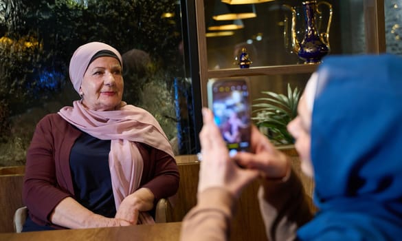In a modern restaurant during the holy month of Ramadan, a woman in a hijab captures a moment with her mother using a smartphone, epitomizing the blending of tradition and technology in familial bonds and capturing cherished memories.