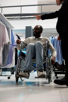 African american man in wheelchair pointing at shirt and asking consultant for advice while shopping in clothing store. Boutique client with disability and employee choosing apparel