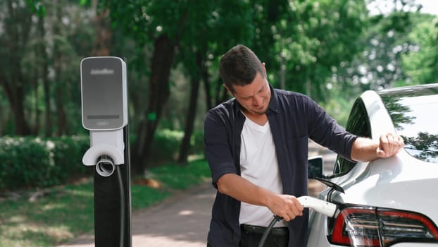 Man recharging battery for electric car during road trip travel EV car in natural forest or national park. Eco friendly travel during vacation and holiday. Exalt