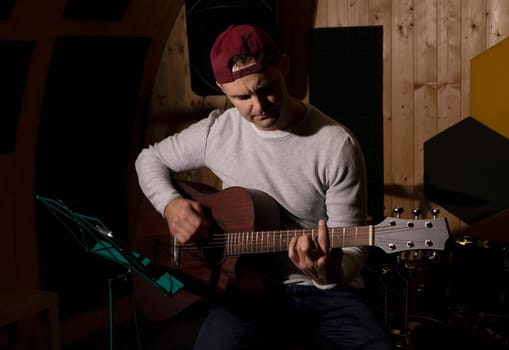 Caucasian man plays guitar, tuning musical instrument in hands, sitting in chair in studio. Darken photo. Male wears casual cloth. Hobby, leisure of creative person, artist. Copy space. Horizontal