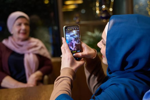 In a modern restaurant during the holy month of Ramadan, a woman in a hijab captures a moment with her mother using a smartphone, epitomizing the blending of tradition and technology in familial bonds and capturing cherished memories.