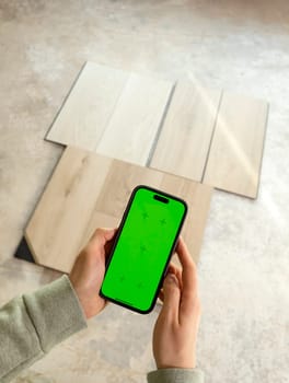 Hands Holding Device with Green Chroma Key Screen above Luxury Vinyl Wood Samples, Laminate On Floor In House. Choosing, Selecting Waterproof Flooring. Home Reconstruction. Vertical Plane.