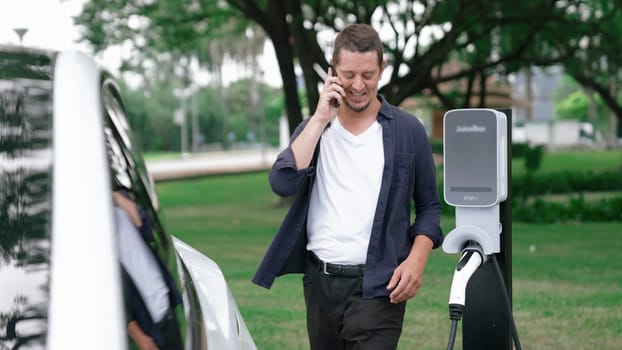 Man recharging battery for electric car during road trip travel EV car in natural forest or national park. Eco friendly travel during vacation and holiday. Exalt