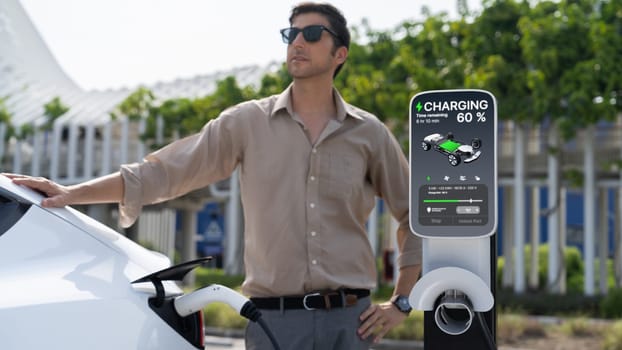 Young man put EV charger to recharge electric car's battery from charging station in city commercial parking lot. Rechargeable EV car for sustainable environmental friendly urban travel. Expedient