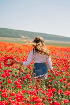 Happy woman in a poppy field in a white shirt and denim skirt with a wreath of poppies on her head posing and enjoying the poppy field